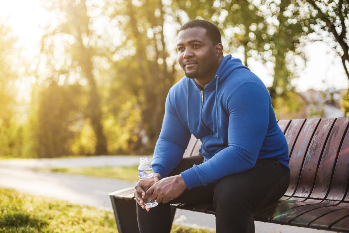 A man sits on a park bench wearing a blue hoodie, holding a water bottle, and enjoying a peaceful moment outdoors in the soft glow of sunlight filtering through the trees.